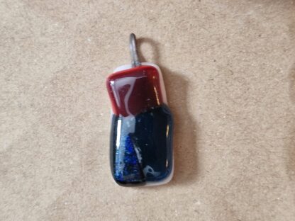 Fusat glassmycke av klarrött, mörkt marinblått och mörkblått glittereffektglas på opakt vitt glas. Infusad ögla av Kanthaltråd. Ca 2.7 x 1.5 cm. 